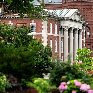 A historic brick building with tall white columns, surrounded by lush greenery and colorful flowers.