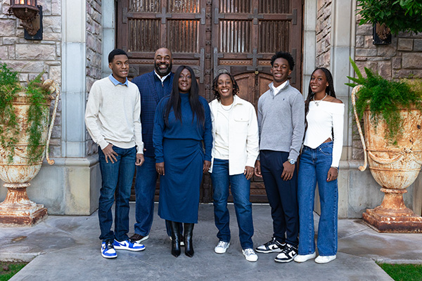 A happy family of six posing in front of their house, smiling towards the camera. They are standing together on the porch, which features large wooden doors and decorative potted plants.