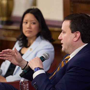 A person in a navy suit and gold-striped tie speaks into a handheld microphone during a panel discussion, gesturing with their left hand. A person in a light blue blazer is seated behind them, slightly out of focus.