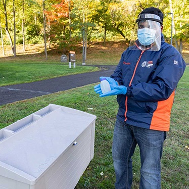 Person in PPE holding a sample bottle beside outdoor equipment on a grassy campus path.