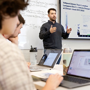Person gesturing while speaking to seated students with laptops, whiteboard and screen behind them.