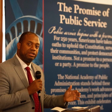 Speaker gesturing while holding microphone near "Promise of Public Service" banner.