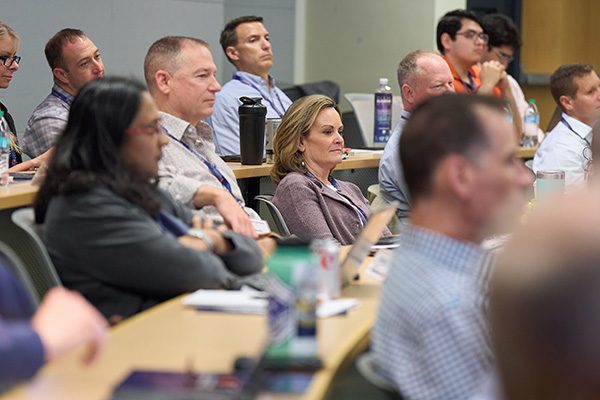 A group of people attentively listening during a conference or lecture. Some have lanyards, notebooks, and bottled water. The setting appears to be a modern conference room.