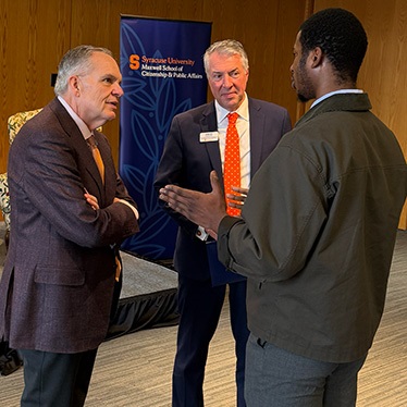 Three individuals are engaged in conversation in front of a Syracuse University Maxwell School banner. One is gesturing with hands while the others listen attentively. They are dressed in business attire.
