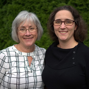Two people smiling together outdoors in front of a green hedge, wearing glasses.
