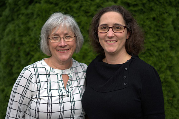 Two people smiling together outdoors in front of a green hedge, wearing glasses.