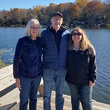 Three adults standing on a wooden dock with a scenic lake and autumn trees in the background.