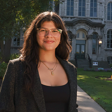 A person with long dark wavy hair and glasses, wearing a black top and gray tweed blazer with a gold necklace, smiling while standing outdoors in front of a stone campus building with a green lawn.
