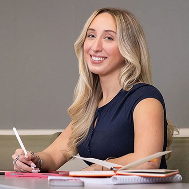 Person sitting at a desk with papers and a digital tablet, smiling and holding a pen, in a professional setting.