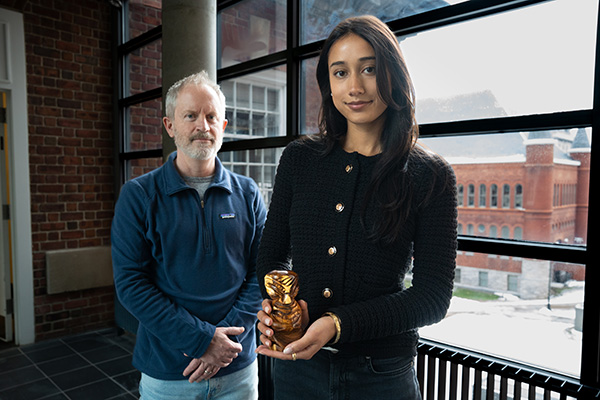 A person holds a small figurine while standing next to another person near large windows overlooking a red brick building.