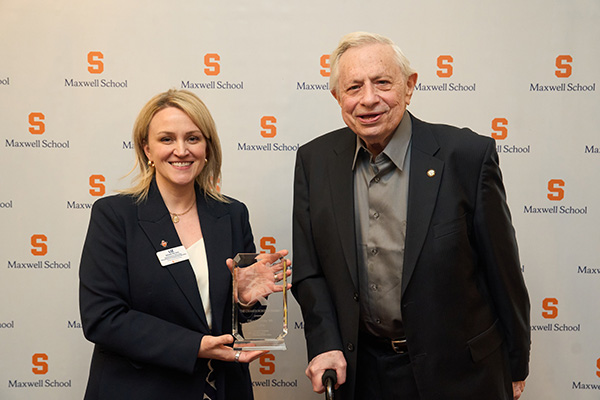 Two individuals at a Syracuse University event, with one holding an award. They are smiling and standing in front of a backdrop with multiple Syracuse University and Maxwell School logos.