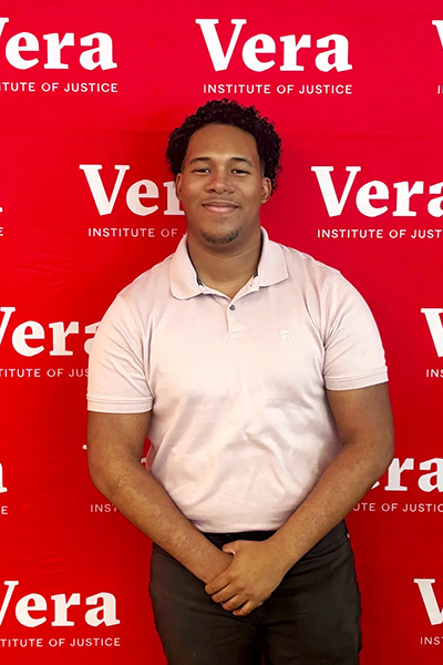 Individual standing in front of a Vera Institute of Justice step-and-repeat banner, wearing a light polo shirt and smiling.