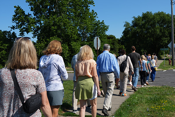 A group walks down a sidewalk past trees on a sunny residential street.