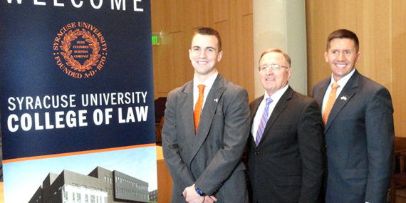 Three individuals stand in front of a Syracuse University College of Law banner, smiling. The setting appears to be an indoor event.