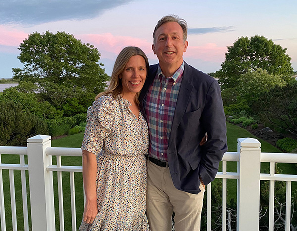 Two people smiling for a photo on a balcony with a scenic sunset in the background.