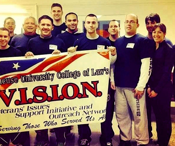 Group of people holding a banner for the Syracuse University College of Law's Veterans' Issues, Support Initiative and Outreach Network (VISION), smiling in a casual indoor setting.