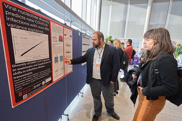 Two individuals are viewing a scientific poster about viral nucleotide diversity and its relation to COVID-19 at a conference. One person is pointing at a graph on the poster.