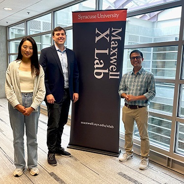 Three individuals standing in front of a Syracuse University Maxwell X Lab banner. They are dressed in business casual attire.