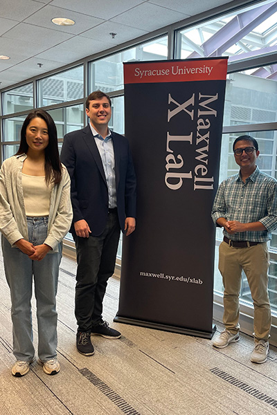Three individuals standing in front of a Syracuse University Maxwell X Lab banner. They are dressed in business casual attire.