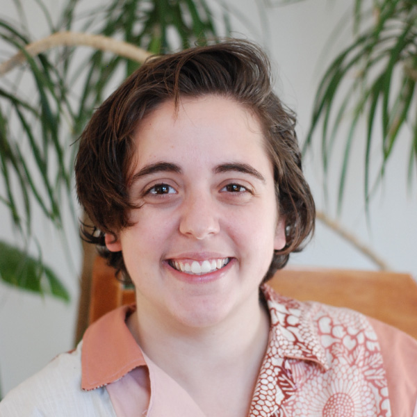 A person with short, dark hair smiling at the camera, wearing a pink shirt and a patterned scarf, set against a blurred indoor background with a plant.