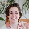 A person with short, dark hair smiling at the camera, wearing a pink shirt and a patterned scarf, set against a blurred indoor background with a plant.