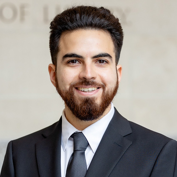 A person with a beard, smiling, wearing a dark suit and tie