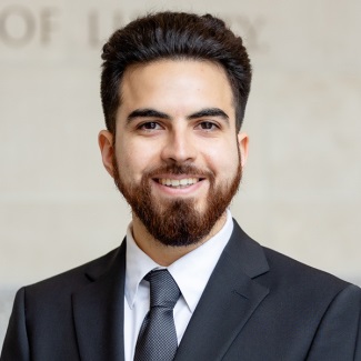 A person with a beard, smiling, wearing a dark suit and tie
