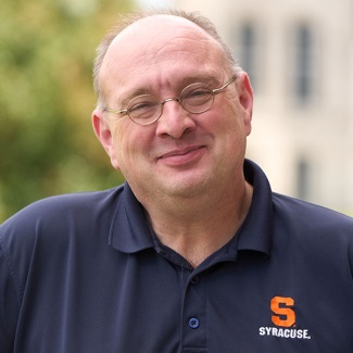 Person smiling and wearing glasses, and a Syracuse University polo shirt. The background is blurred with green foliage.