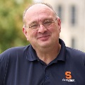 Person smiling and wearing glasses, and a Syracuse University polo shirt. The background is blurred with green foliage.