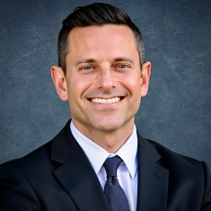 A person with short dark hair and light eyes, smiling broadly at the camera. They are wearing a dark suit jacket, light blue dress shirt, and a navy patterned tie. The background is a muted gray-blue textured backdrop.
