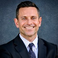 A person with short dark hair and light eyes, smiling broadly at the camera. They are wearing a dark suit jacket, light blue dress shirt, and a navy patterned tie. The background is a muted gray-blue textured backdrop.