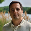 A person wearing a cream polo shirt with a lake and tall reeds in the background.
