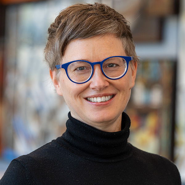 Portrait of a smiling individual with short hair, wearing blue round glasses and a black turtleneck, standing in a room with blurred paintings in the background.