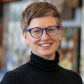 Portrait of a smiling individual with short hair, wearing blue round glasses and a black turtleneck, standing in a room with blurred paintings in the background.