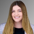 Professional headshot of a smiling individual with long blond hair, wearing a tweed jacket against a neutral background.