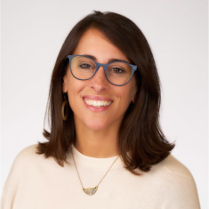 A smiling person with dark brown hair and glasses wearing a light pink top and a gold necklace.