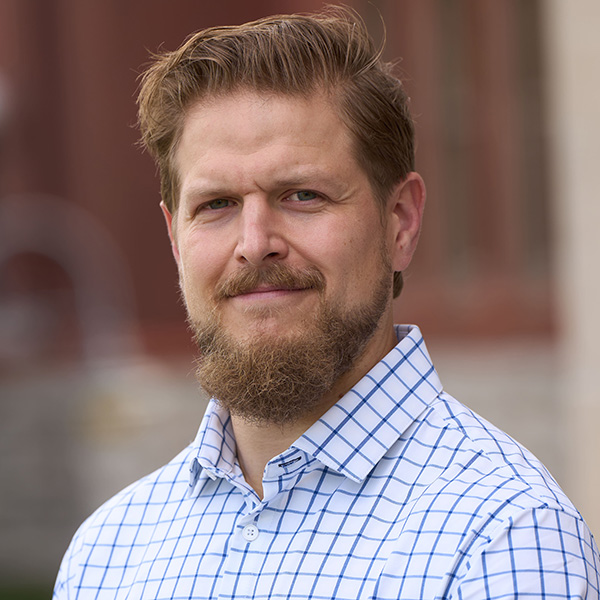 Portrait of a smiling person with a beard, wearing a blue and white checked shirt. Background suggests an outdoor setting with blurred architecture.