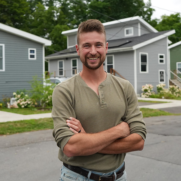 A person stands with arms crossed on a street lined with tiny homes.