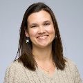 Portrait of a smiling person wearing a beige sweater and hexagonal earrings, set against a plain gray background.
