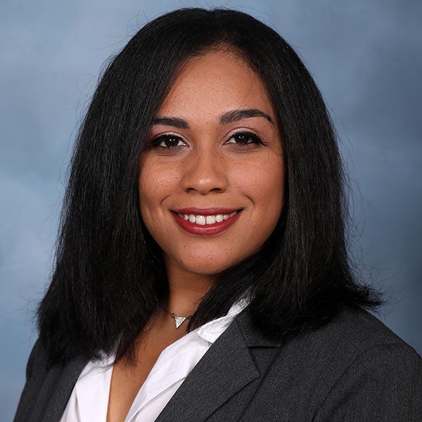 A person smiling against a soft blue-gray background. They are wearing a gray blazer over a white blouse and a delicate silver necklace.