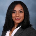 A person smiling against a soft blue-gray background. They are wearing a gray blazer over a white blouse and a delicate silver necklace.