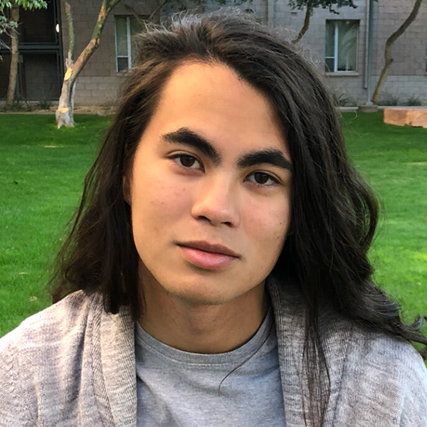 Close-up portrait of a person with long dark hair, wearing a gray t-shirt and light gray cardigan, sitting outdoors with greenery and a building background.