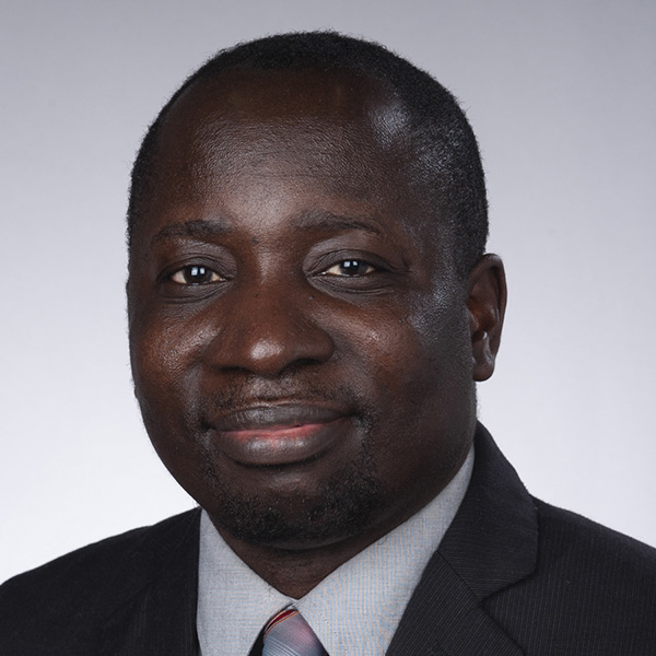 A person in a dark suit jacket, light dress shirt, and striped tie smiles at the camera against a light gray studio background
