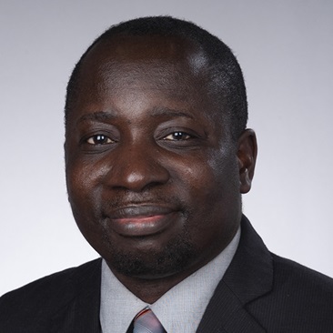A person in a dark suit jacket, light dress shirt, and striped tie smiles at the camera against a light gray studio background