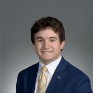 A person with short brown hair wearing a blue suit, white shirt and yellow tie.