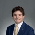 A person with short brown hair wearing a blue suit, white shirt and yellow tie.