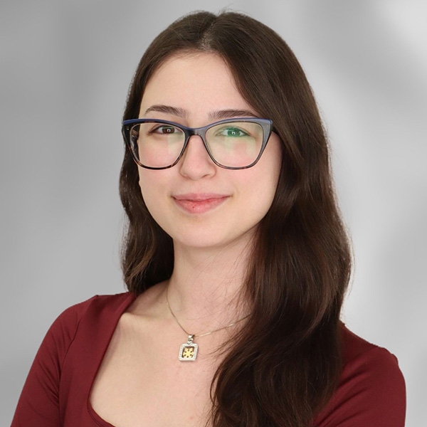 A person with long brown hair wearing a reddish top, glasses and a necklace.