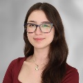 A person with long brown hair wearing a reddish top, glasses and a necklace.
