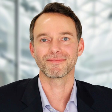 A professional portrait of an individual in a dark suit and blue shirt, featuring a subtle smile and set against a blurred architectural background.