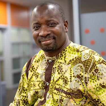 A person smiling, wearing a patterned yellow and brown traditional shirt.